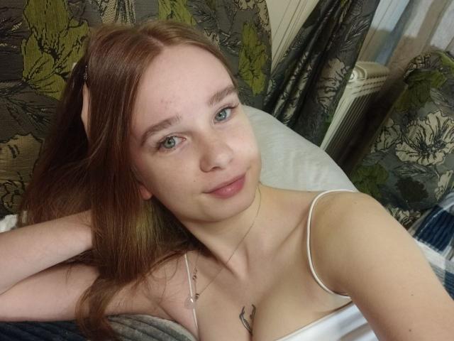 StaceyKiss has red hair, green eyes, and a small tattoo on her chest. She's lying on a bed, wearing a white spaghetti strap top, smiling at the camera.