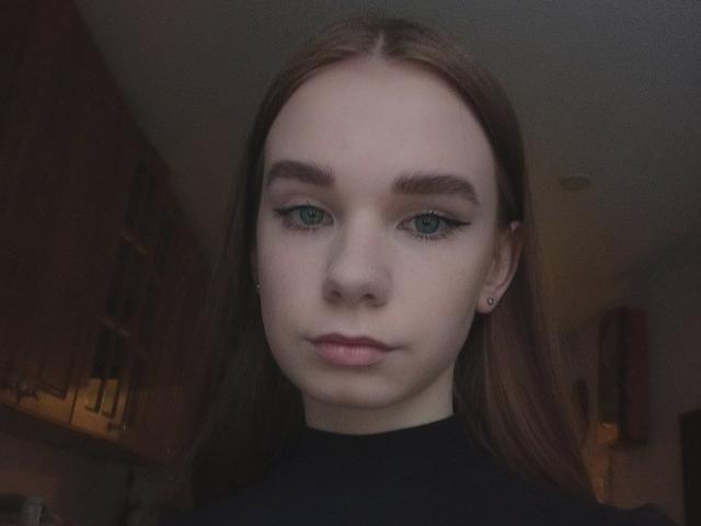 StaceyKiss has pale skin, green eyes, and long brown hair, wearing a black top, with subtle makeup and dark eyebrows. She's looking straight at the camera with a neutral expression.
