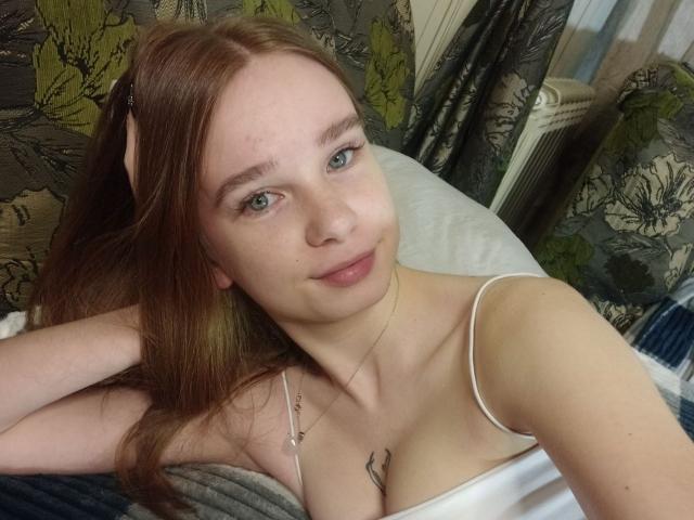 StaceyKiss with red hair, green eyes, and small tits, wearing a white spaghetti strap top, lying on a bed, smiling slightly, with a necklace and tattoo on her chest.