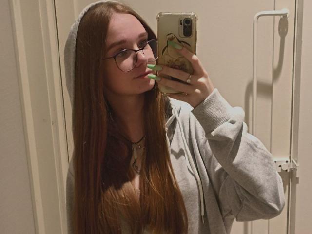 StaceyKiss with long brown hair, glasses, green nails, gray hoodie, and beanie, taking a mirror selfie with a phone, showing off her cute face and sexy vibe.