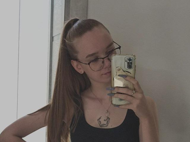 StaceyKiss with long brown ponytail, glasses, black tank top, holding phone, grey nails, necklace with crescent moon, mirror selfie, standing by window, neutral expression.