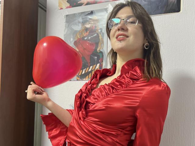 IsleBelle with glasses, red satin blouse, holding a red heart balloon, smiling, brown hair, small nose ring, wall with anime posters, white textured wall, casual photo.