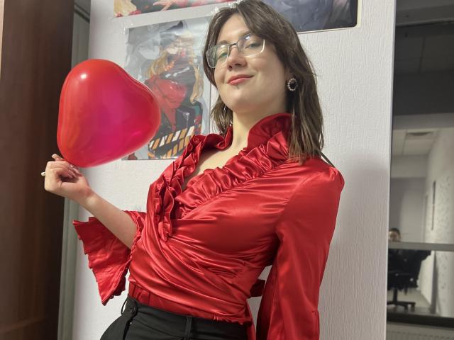 IsleBelle with glasses, red ruffled satin shirt, holding pink heart balloon, black pants, medium tits, slightly wavy brown hair, white wall, partial mirror reflection, confident smile.