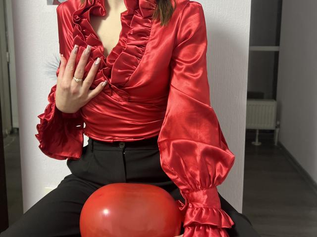 IsleBelle in shiny red ruffled blouse, black pants, hand on chest, sitting with red balloon, showing tits, wearing gold ring, indoors, photo, realistic, sexy.