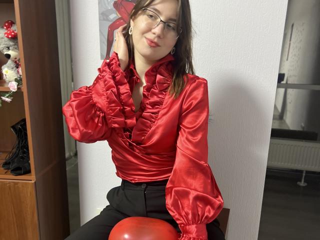 IsleBelle with red ruffled satin shirt, black pants, and glasses, sitting on a red balloon, hand in hair, smiling, medium tits, fair skin, brown hair, red lipstick.