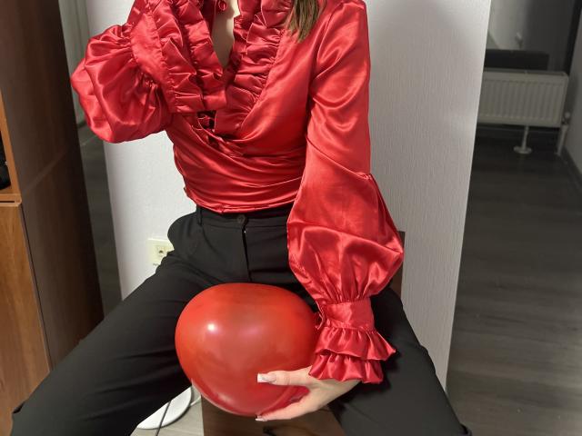 IsleBelle in a shiny red ruffled blouse, holding a red ball between her black pants, sitting with her hand on her chest, wearing a red necklace.