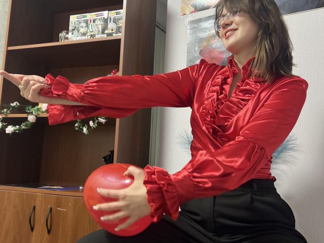 IsleBelle with shoulder-length brown hair, wearing a red ruffled shirt and black pants, holding a red balloon while pointing with her other hand, smiling.