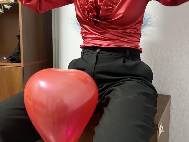IsleBelle's red satin top, black pants, and shiny red balloon between her legs. Her top is tied at the chest, highlighting her tits. She's sitting on a brown box.