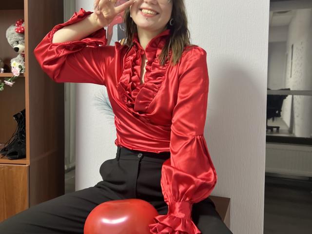 IsleBelle in a shiny red ruffled blouse, black pants, and glasses, smiling with a peace sign, sitting on a red balloon. Her tits are covered by the blouse.
