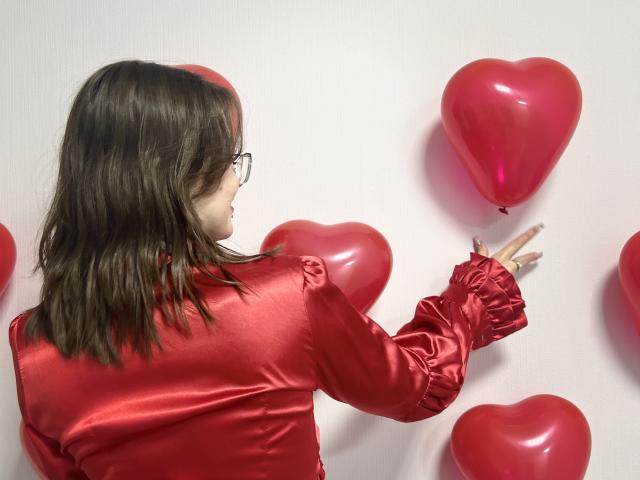 IsleBelle with red satin shirt, brown hair, glasses, reaching for pink heart balloons, wearing shiny red sleeves, back to camera, white wall, pink heart balloons around.