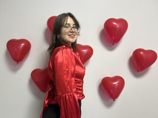 IsleBelle with red satin blouse, black pants, glasses, and earrings, smiling at camera, surrounded by red heart balloons, standing against white wall.