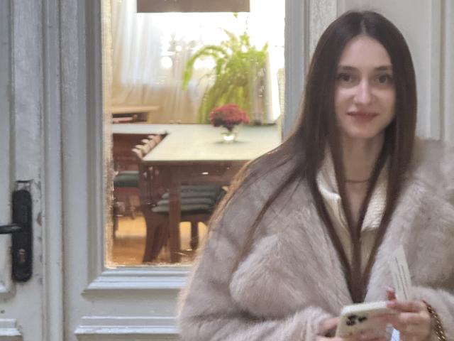 LaraSweetyHot met lang bruin haar, gekleed in een pluizige beige bontjas, met een telefoon in haar hand, staand bij een raam met een lichte glimlach, en een schattige uitstraling..
