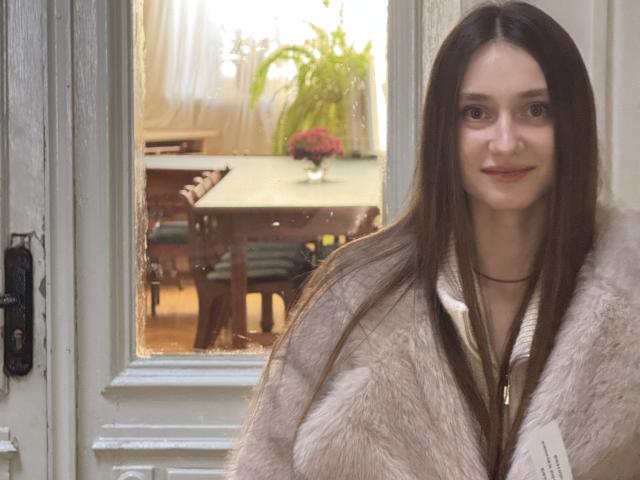 LaraSweetyHot, con lunghi capelli castani, sorridente, indossa un cappotto beige in pelliccia, è in piedi davanti a una finestra con cornice bianca, tiene in mano una busta bianca, appare carina e sexy..