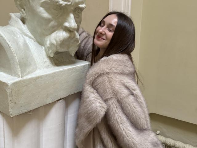 LaraSweetyHot, avec de longs cheveux bruns, souriante, portant un manteau gris en fourrure moelleuse, appuyée contre un buste en marbre blanc dans une pièce..