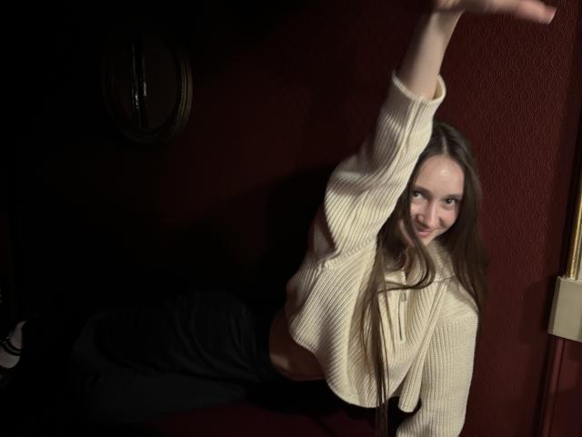 LaraSweetyHot met lang bruin haar, lachend, gekleed in een beige gebreide crop top, zwarte broek, met één arm omhoog, waarbij haar getoonde buik zichtbaar is, in gedimd licht..