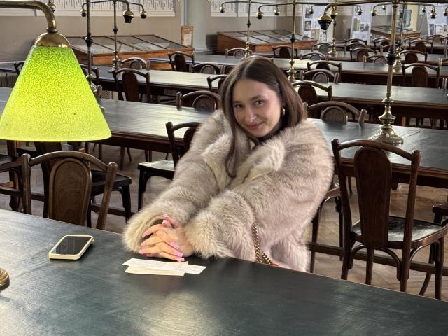 LaraSweetyHot with tan skin, brown hair, and a fluffy gray fur coat, sitting at a green table, hands on it, green lamp, phone, and white card in front.