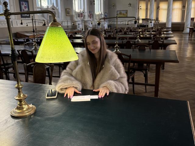 LaraSweetyHot met lang bruin haar, gekleed in een pluizige beige bontjas, zittend aan een groene tafel, met papieren in haar handen, onder een groene glazen lamp, in een houten kamer..