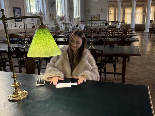 LaraSweetyHot, avec ses longs cheveux bruns, portant un manteau beige en fourrure, est assise à une table verte, touchant une feuille de papier blanche, avec une lampe verte texturée devant elle..