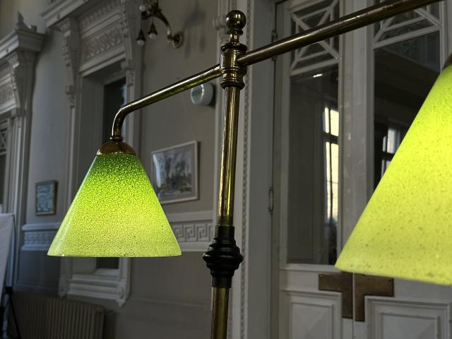 Golden brass floor lamp with two green, textured, cone-shaped boobs, aka lamps, hanging from a curved arm, in a classy, old-school room with white walls and detailed trim.