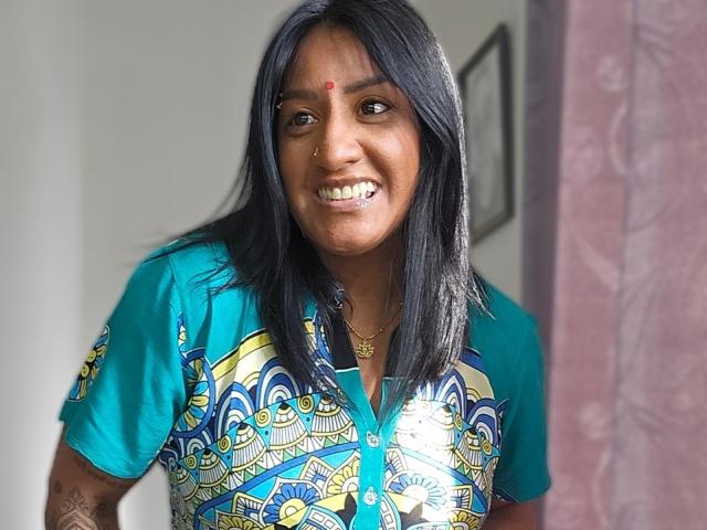 Nahirr, brown-skinned woman with black hair, small red dot on forehead, smiling, blue patterned shirt, gold necklace, cigarette in mouth, right arm tattoo, pink curtain.