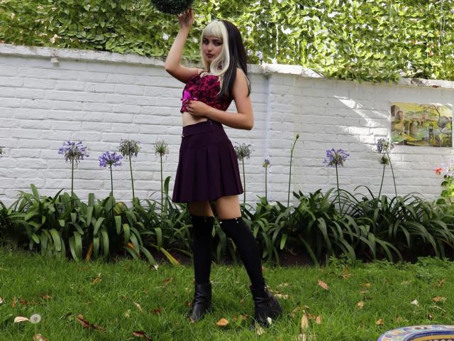 AngelBlakee with dyed black and white hair, purple skirt, black knee socks, and pink floral top, holding a black bra, standing on grass, showing her small tits.