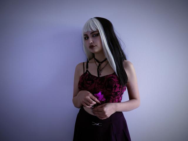 AngelBlakee with white and black hair, dark lipstick, wearing a pink and black lace top, holding a purple flower, looking at camera with a slightly sad expression.