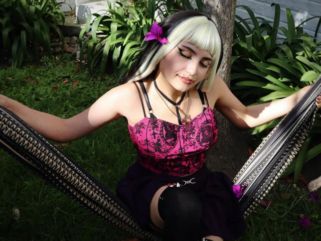 AngelBlakee with pale skin, green-black hair, pink-purple top, black straps, black skirt, and black thigh-highs, sitting on a mirror, grass, and plant background, looking down.