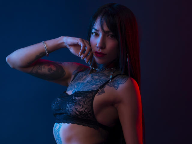 KaydeEbexley with tattoos, wearing black lace bra, arm raised, red and blue lighting, looking at camera, red lipstick, chain necklace, black hair, slim, visible tattoos on chest and arm.