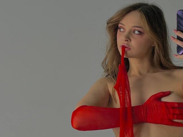 MayxFlower with long brown hair, red glossy gloves, and red strap pulling from her mouth, looking to the side, holding a phone. Her tits are covered by her hand.