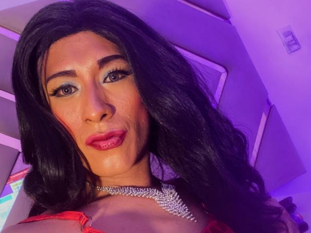 ThomasParker69 has long black hair, blue eye makeup, red lipstick, and a sparkly necklace. Purple lighting highlights their face, showing a confident, sexy expression. Boobs visible in red top.