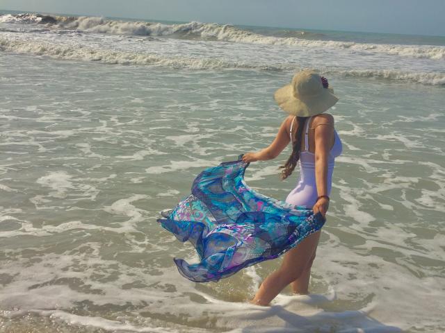 DanesaCronin with tan skin, wearing a white dress and sunhat, holding a blue and purple patterned scarf, standing in shallow ocean water with waves around her.