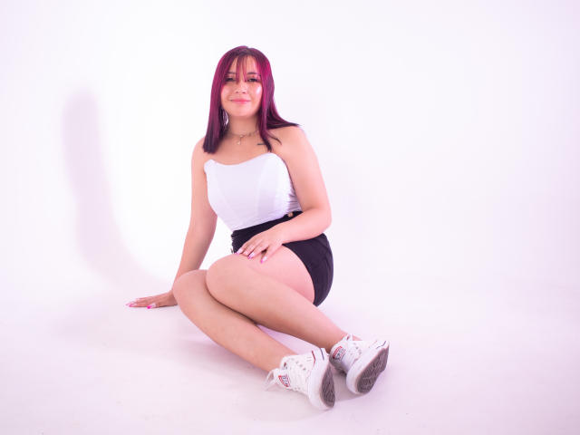 AriaBliss, with purple hair, sits on the floor wearing a white strapless top, black shorts, and white sneakers, showing her ass and smiling.