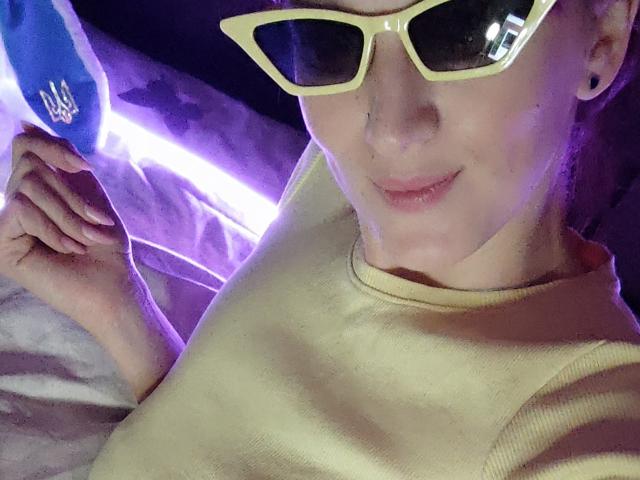 FavoriteHare portant des lunettes de soleil jaunes et une chemise jaune, souriant avec un petit anneau de nez, levant la main, des lumières violettes et bleues en arrière-plan..