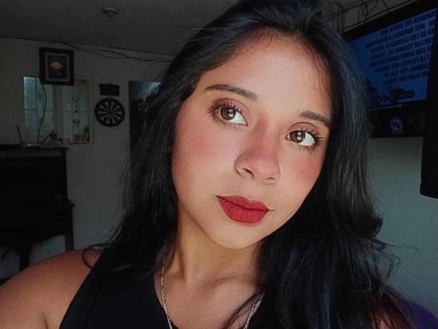 ChloeCanela has dark, wavy hair, brown eyes, and red lipstick. She's wearing a black top and a silver necklace, with a serious, slightly pouty expression on her face.