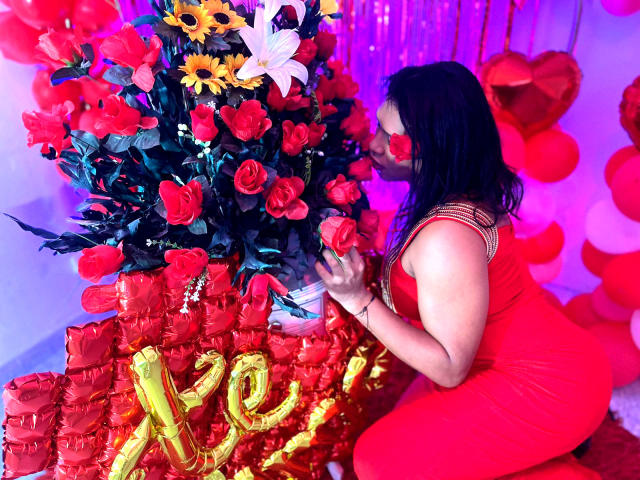 ZahorySexy with long dark hair, red dress, and red mask, kneeling, touching bright red and yellow rose bouquet, surrounded by red balloons and gold "XO" letters.