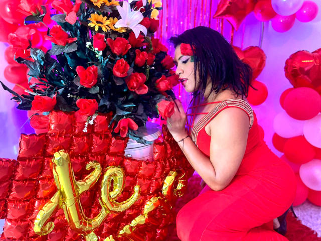 ZahorySexy in a red dress with gold trim, eating a red rose, kneeling by a red rose bouquet, surrounded by red balloons, and a golden "A" balloon.