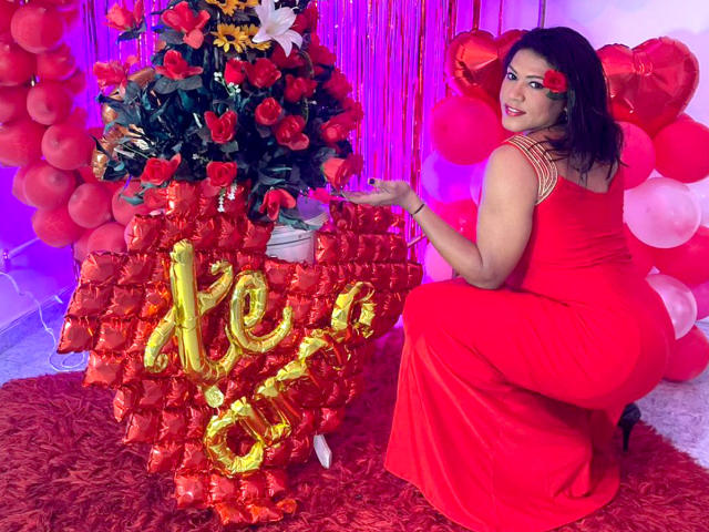 ZahorySexy, in a red dress, kneels beside a rose-adorned tree with a gold "20" balloon, red balloons, and a red floral rug, with a red rose in her hair.