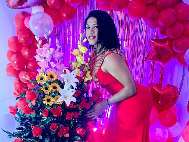 ZahorySexy with black curly hair, red dress, and red necklace, leaning on a flower and balloon arch, showing her ass, with colorful flowers and red balloons.
