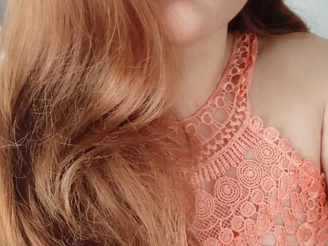 Le visage d'AnaGinger est partiellement visible, avec un accent sur son soutien-gorge en dentelle orange et ses longs cheveux ondulés, d'une nuance de roux-brun, qui recouvrent son épaule droite. Sa peau est claire..