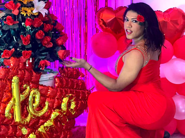 ZahorySexy, in a red dress, touches red roses. She has a rose in her hair, a cute ass, and is smiling. Balloons and pink lights are in the background.