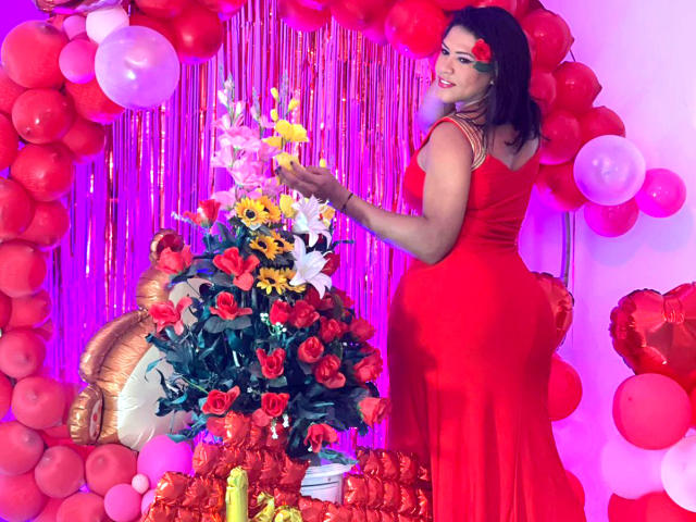 ZahorySexy in a red dress, holding flowers, standing beside a flower arrangement with red roses and yellow flowers, surrounded by red and white balloons, looking back.