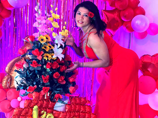 ZahorySexy in a red dress with a flower in her hair, leaning on a colorful bouquet of roses and sunflowers, surrounded by red and pink balloons.