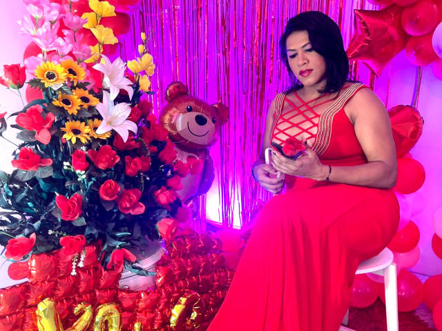 ZahorySexy, a curvy woman with black hair, red dress, and visible cleavage, sits holding a small red box, next to a teddy bear and vibrant red flower arrangement.