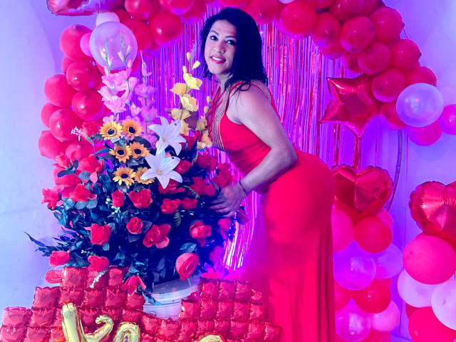 ZahorySexy, with red dress, leans on flower arrangement of sunflowers, roses, and lilies. Her ass is prominent, surrounded by red balloons and heart-shaped decorations. Purple light.