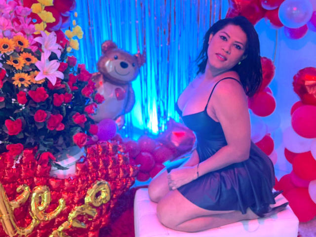 ZahorySexy, with tits showing in a sheer black dress, kneeling on a white cube, surrounded by red roses, yellow lilies, blue lights, and a teddy bear.
