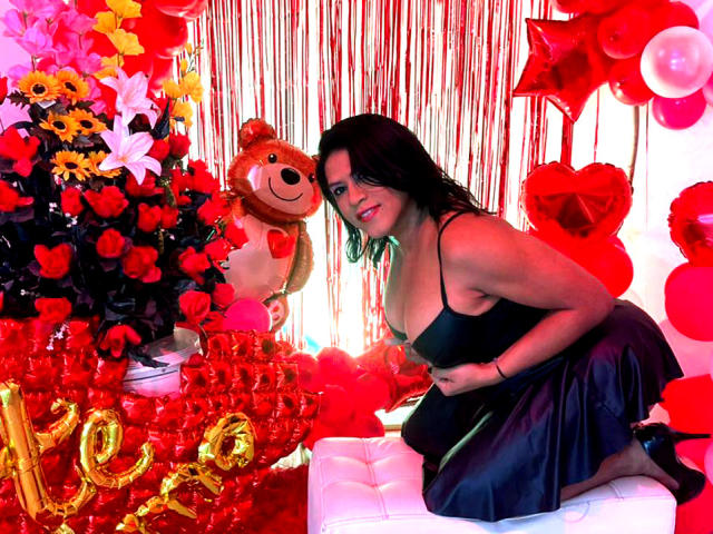 ZahorySexy with big tits, black dress, kneeling on white bench, surrounded by red roses, balloons, and a teddy bear, smiling at the camera in a colorful, festive setting.
