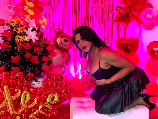 ZahorySexy, with dark hair and red lipstick, kneels in a black satin dress, next to a teddy bear and red rose bouquet, surrounded by pink and red balloons.