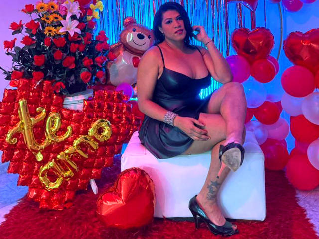 ZahorySexy, curvy with big boobs, in a black sheer dress, sitting on a white cube, legs crossed, black heels, tattoo on right calf, red and blue background.