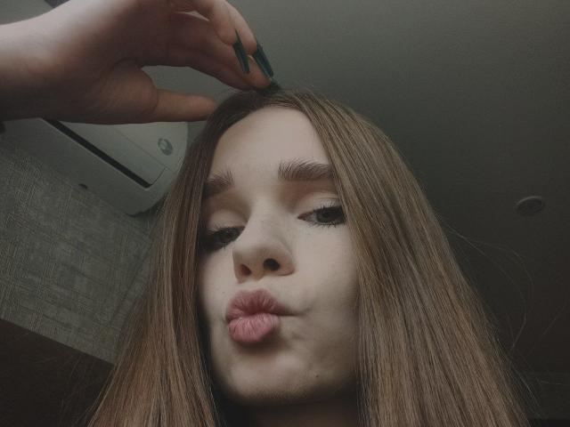 StaceyKiss with long brown hair, pouty pink lips, and a hand holding her hair up, showing off her green nail polish. Her eyes are half-closed.