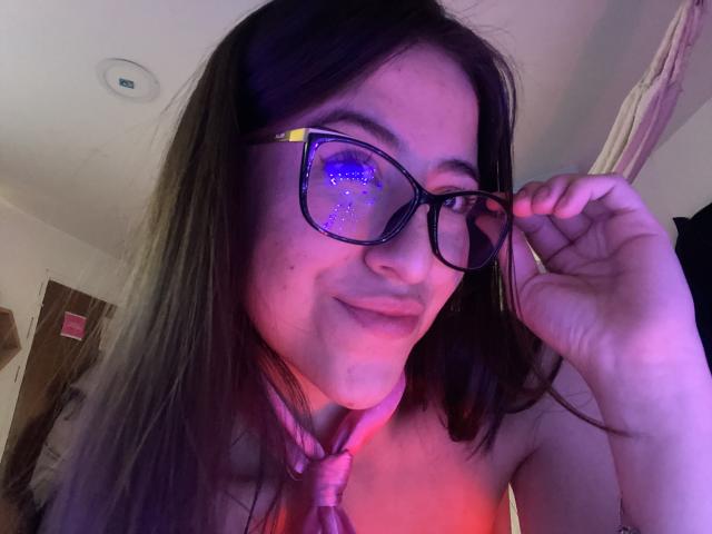 KatiaRogers with purple-lit glasses, holding frame, wearing pink tie, smiling, brown hair, fair skin, purple and pink lighting, in a selfie, showing her face and upper chest.